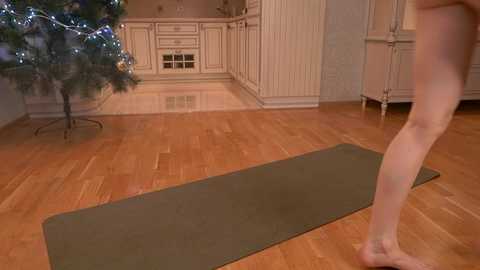 Media: A video of a light-skinned, nude person standing on a yoga mat in a cozy, wooden-floored kitchen with cream-colored cabinets and a Christmas tree in the corner.