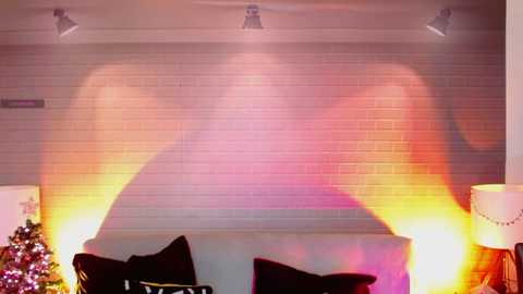 Media: Video of a modern living room with a textured, light gray brick wall displaying a colorful, abstract light projection. A white sofa with black pillows and a decorative Christmas tree with fairy lights are visible.