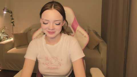 Media: A video of a young woman with fair skin, brown hair, and a white T-shirt, seated in a pink gaming chair, indoors with beige furniture and a potted plant in the background.