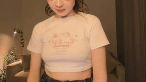 Media: Video of a young East Asian woman with fair skin, wearing a white crop top with a bunny design and blue jeans, in a dimly lit room with beige walls and a lamp.