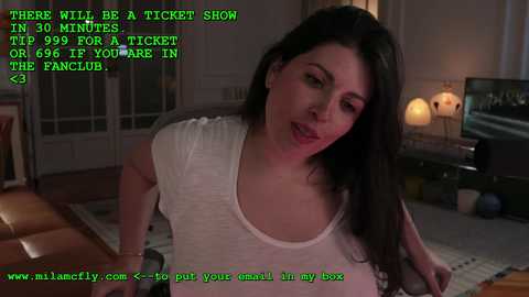 Media: A video of a woman in a white tank top with medium breasts, dark hair, and a serious expression, taken in a dimly lit living room with a green computer screen overlay.