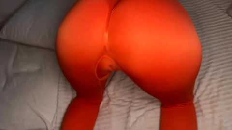 Media: Video of a person with light skin, wearing tight, bright orange leggings, lying face down on a striped bed, emphasizing their buttocks and thighs.