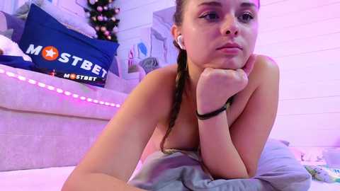 Media: Video of a young, light-skinned woman with a slender build, topless, lying on a bed with a grey blanket. She has long brown hair braided, wearing white earbuds, looking contemplative. Background shows a Christmas tree, pillows, and a \"MOSTBET\" pillow.