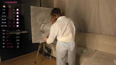 Media: A video of a woman with long brown hair painting a nude figure on an easel in a dimly lit room. She wears a white cardigan and light blue pants.