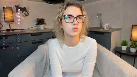 Media: Video of a young woman with curly blonde hair, wearing black-rimmed glasses and a white long-sleeved top, seated in a gray armchair in a modern kitchen.