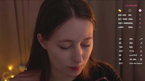 Video of a young woman with fair skin and long brown hair, wearing a dark top, gazing downward, set in a dimly lit room with a cozy ambiance.