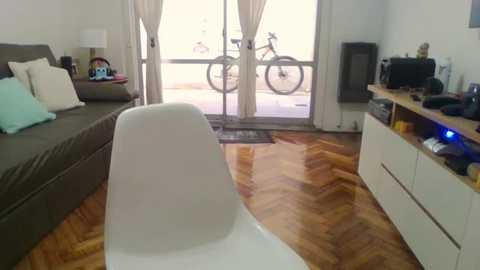 Media: Video of a modern living room with a grey sectional sofa, white chair, wooden herringbone floor, white curtains, and two bicycles outside a glass door.