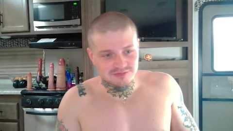 Media: Video of a bald, light-skinned man with a short beard and tattoos on his arms and chest, smiling in a modern kitchen with stainless steel appliances, microwave, and a knife block.