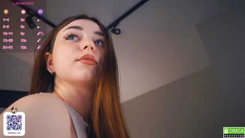 Media: Video of a young Caucasian woman with fair skin and long, straight brown hair, wearing a white sleeveless top, looking upward, in a dimly lit room with a calendar and a QR code visible.