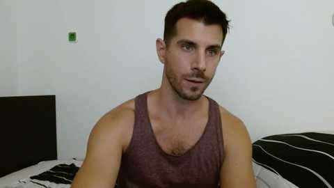 Media: Video of a muscular, light-skinned man with short dark hair, wearing a sleeveless gray tank top, sitting on a bed with a black and white striped pillow.