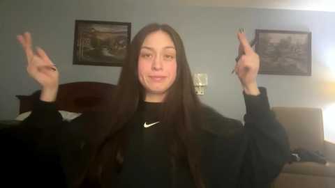 Media: Video of a young woman with long, straight brown hair, wearing a black top, smiling, and making a peace sign with both hands in a dimly lit bedroom with two framed landscapes on the wall.