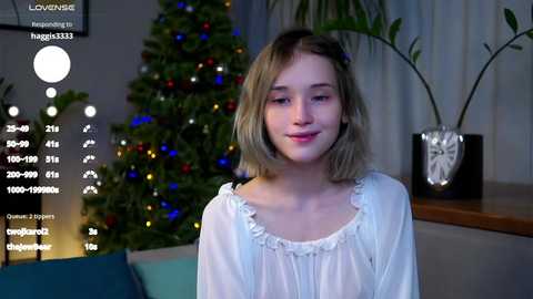 Video of a young, fair-skinned girl with shoulder-length blonde hair, wearing a white blouse, sitting indoors beside a decorated Christmas tree and potted plants.