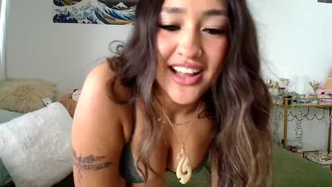 Media: Video of a smiling, curvy Asian woman with long, wavy brown hair, wearing a green top, gold necklace, and nose ring, in a cozy, cluttered room with a white wall, blue and white artwork, and a beige bed.