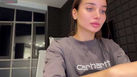 Media: Video of a young woman with fair skin, blue eyes, and straight brown hair, wearing a gray \"Gymnastik\" shirt, sitting in a modern indoor setting with dark brick walls and large windows.