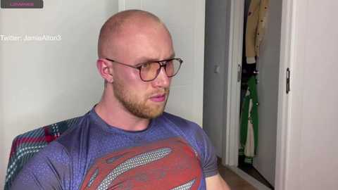 Media: Video of a bald, bearded, fair-skinned man with glasses wearing a Superman T-shirt, standing in a closet with hanging clothes, against a white background.