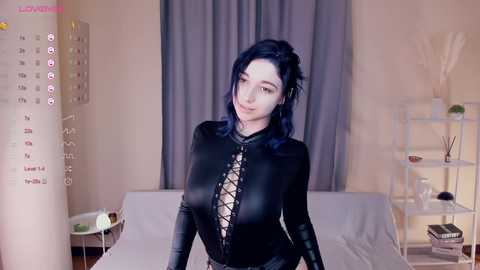 Media: Video of a pale-skinned, dark-haired woman in a black, form-fitting latex bodysuit with a plunging neckline, standing in a modern bedroom with a bed, shelves, and a calendar.