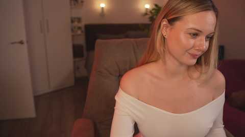 Media: Video of a smiling, light-skinned woman with long blonde hair, wearing an off-shoulder white top, sitting indoors with beige furniture, warm lighting, and a blurred background.