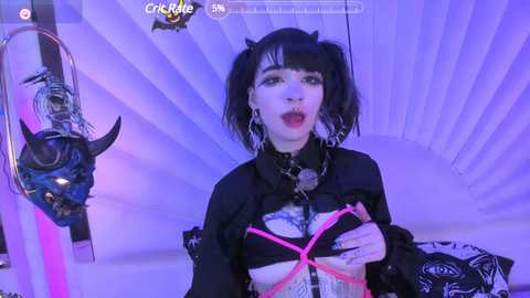 Media: Video of a pale-skinned woman with black hair in pigtails, wearing a black crop top with pink straps and a metal chain, standing in a room with a purple, striped background.