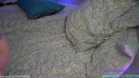 Media: Video of a person, possibly male, with a grey, textured sweater, lying on a bed with a teal pillow and a blue light illuminating the room, captured by a webcam.