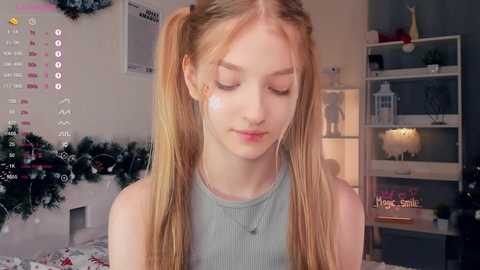 Media: Video of a fair-skinned, blonde-haired young woman with pigtails, wearing a light gray top, in a cozy, decorated room with a Christmas tree, calendar, and shelves.