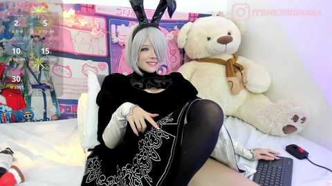 Media: Video of a smiling woman in a black, white, and red cosplay outfit with bunny ears, sitting on a bed with a teddy bear. Background features a colorful anime poster and a computer screen.