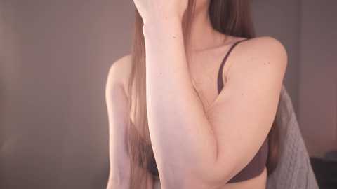 Media: Video of a fair-skinned woman with long brown hair, wearing a dark bra, partially covering her face with her right arm, against a blurred background.