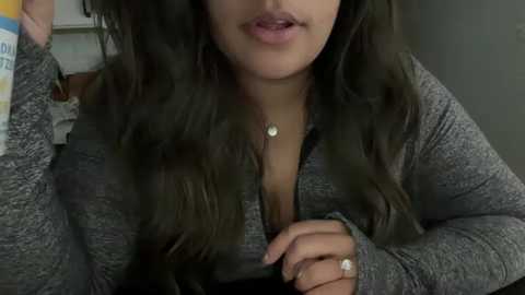 Media: Video of a woman with long, dark hair, wearing a grey cardigan and a pendant necklace, lying on a bed with a striped pillow, partially visible.