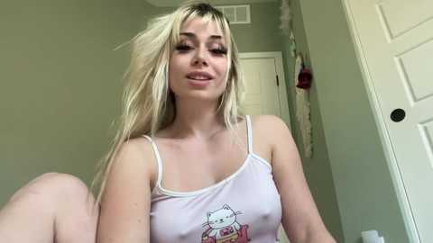 Media: A video of a blonde woman with fair skin, wearing a white tank top with a Hello Kitty design, sitting in a room with light green walls and a white door.