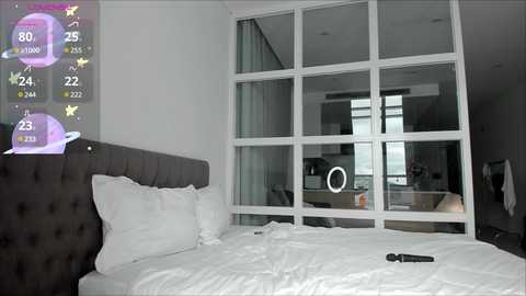 Media: A modern, minimalist bedroom with a dark gray tufted headboard, white bedding, and a large window with a circular light fixture. A smartphone sits on the bed.