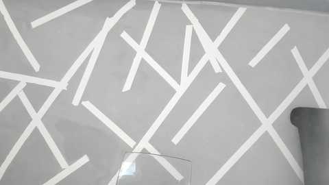 Media: A video of a modern, minimalist room with a white wall covered in white painter's tape forming intersecting, crisscrossing patterns. A gray armchair and a clear glass object are visible in the foreground.