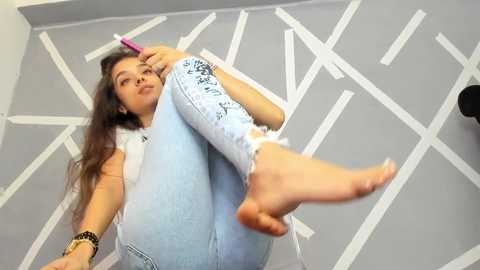 Media: Video of a young woman with long, wavy brown hair, wearing light blue distressed jeans and a white tank top, sitting on a floor with white geometric patterns.