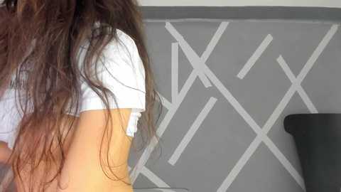 Media: Video of a person with long, wavy brown hair, wearing a white shirt, standing in a room with a gray wall featuring white geometric patterns.