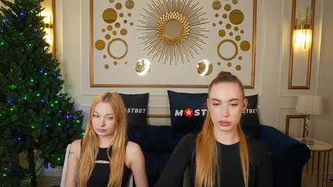 Media: Video of two blonde, fair-skinned women in black dresses, standing side by side in a stylish, ornate room with a sunburst mirror, a Christmas tree, and modern d\u00e9cor.