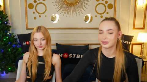 Media: Video of two young women with long blonde hair, dressed in black crop tops, seated on a sofa. Background includes a Christmas tree, golden sunburst mirror, and \"MST3K\" pillows.