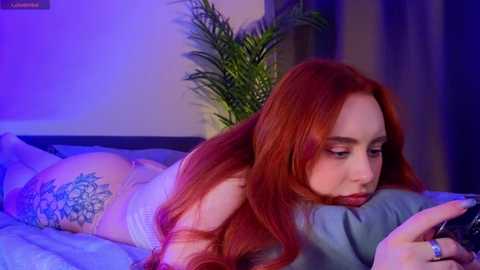 Media: Video of a fair-skinned woman with long, red hair lying on her stomach on a bed, wearing white lingerie, holding a phone, with a large floral tattoo on her hip, in a dimly lit room.