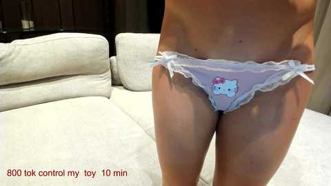 Media: Video of a light-skinned woman's torso, wearing sheer, pink Hello Kitty panties with lace trim, standing in a modern living room with beige couches.