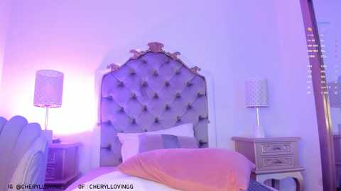 Media: Video of a cozy, modern bedroom with a tufted gray headboard, a light wooden nightstand, and a white lamp with a patterned shade. A peach-colored throw blanket drapes over the bed, and a mirror reflects the soft purple lighting.