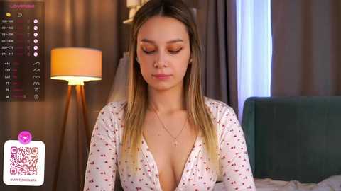 Media: Video of a young woman with light skin and long blonde hair, wearing a low-cut white blouse with red polka dots. She sits in a dimly lit bedroom with a green upholstered headboard.