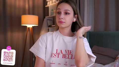 Media: Video of a young woman with long brown hair, wearing a white T-shirt with red text, seated in a dimly lit room with a tall floor lamp, and a teal couch in the background.