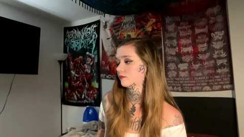 Media: Video of a young woman with long blonde hair, multiple tattoos, and dark makeup, standing in a dimly lit room with heavy metal posters and a blue lamp.