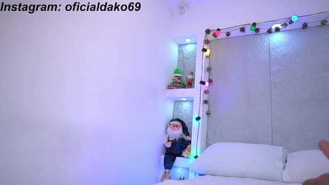 Media: Video of a cozy bedroom with white walls and a mirror adorned with colorful Christmas lights. A small elf figurine stands on a white shelf, and a white bed with pillows is visible.