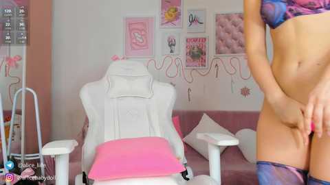 Media: Video of a light-skinned woman in a pink bra and panties, standing in a pink room with white chairs and heart-shaped pillows.