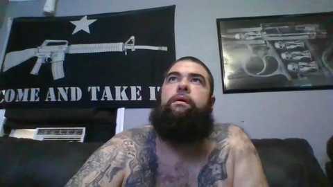 Media: Video of a bearded man with tattoos on his chest and arms, sitting on a couch in a room with a \"Come and Take It\" flag and a gun poster on the wall.