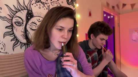 Media: Video of two young people, a brunette woman and a man with short hair, smoking a hookah in a cozy, dimly lit room with a sun and moon tapestry and string lights.