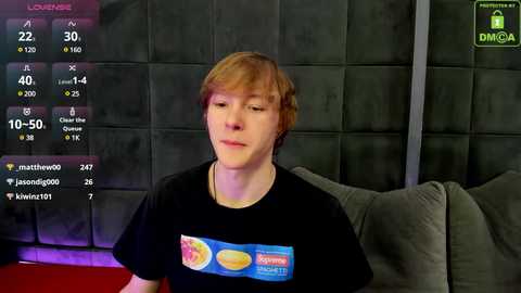 Media: Video of a young man with light skin, light brown hair, and a black t-shirt featuring a \"Dunkin' Donuts\" logo. He sits on a gray couch in a dimly lit room with a dark green, quilted wall.