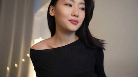 Media: Video of a young East Asian woman with smooth skin, straight black hair, and subtle makeup. She wears a black off-shoulder top. Background features soft, blurred lighting and a white curtain.