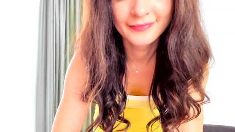 Media: Video of a smiling young woman with long, wavy brown hair, wearing a yellow tank top, standing indoors with light-colored curtains in the background.