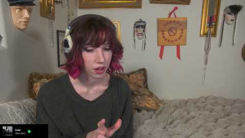 Media: Video of a pale, androgynous person with short, wavy purple hair wearing a dark sweater and large headphones, in a cozy room with framed art, a plush bed, and a doll head.