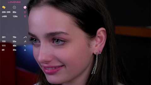 Media: A close-up video of a young woman with fair skin, dark hair, and green eyes, smiling softly. She wears minimal makeup and long, dangling earrings. The background is blurred, featuring a red and black setting.