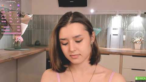 Media: Video of a young woman with medium-length brown hair and fair skin, wearing a light pink spaghetti-strap top, in a modern kitchen with tiled backsplash and stainless steel appliances.